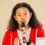 マイクを持って話している男性の写真。長い髪を後ろに流し、赤いパーカーと白地にテキストが印刷されたTシャツを着ている。