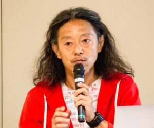 マイクを持って話している男性の写真。長い髪を後ろに流し、赤いパーカーと白地にテキストが印刷されたTシャツを着ている。