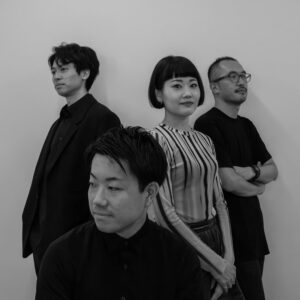 モノクロ写真で、4人の大人が立っている様子を映している。前列には黒いシャツを着た男性が座っており、後ろの列にはひとりの男性とひとりの女性が立っています。一番後ろには腕を組んだ男性が一人立っている。女性のみカメラの方を向いており、女性を除いて全員が黒い服を着用している。女性はボーダーの入った白と黒のストライプのトップスに黒いボトムスを合わせている。