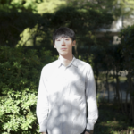 白いストライプのシャツを着た若い男性が、自然の中でカメラを真正面から見つめて立っている写真。