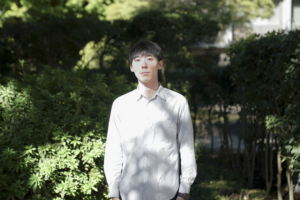 白いストライプのシャツを着た若い男性が、自然の中でカメラを真正面から見つめて立っている写真。