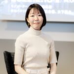 白いタートルネックのトップスを着た女性が、明るい部屋の中でカメラに向かって微笑んでいる写真