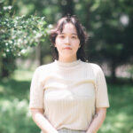 柔らかなクリーム色のトップスを着た女性。明るく緑の木々に囲まれた場所に立っている写真。