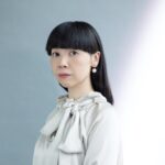 襟元に大きなリボンがあしらわれたオフホワイトのブラウスを着た、肩までの真っ直ぐな黒髪を持つ女性