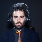 実験的な装置を頭に装着した男性の写真。スーツとネクタイを着用し、真剣な表情でカメラを見つめている。
