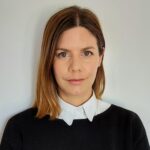 アレクシア・アヒレオス氏の写真。ブラウンとブロンド髪の、白い襟のついた黒いセーターを着ている女性。白い壁を背景に、正面を向いて穏やかな表情をしている。