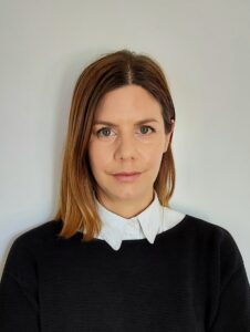 アレクシア・アヒレオス氏の写真。ブラウンとブロンド髪の、白い襟のついた黒いセーターを着ている女性。白い壁を背景に、正面を向いて穏やかな表情をしている。