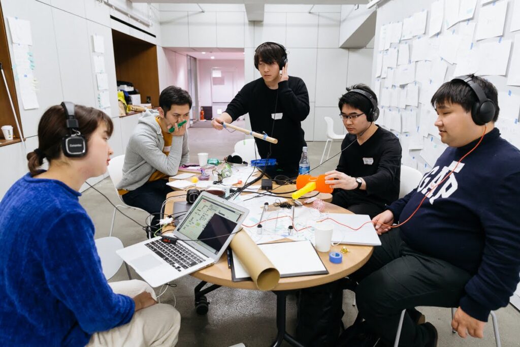 ヘッドホンを着けた5人の男女が円形のテーブルを囲んで、電子機器やノートパソコンを使いながら作業している。テーブルの上には紙、ペン、ケーブル、工具などが広がっており、集中して議論や作業を行っている様子が伺える。背景には白い壁に多くの紙が貼られており、ミーティングルームのような環境が見える。