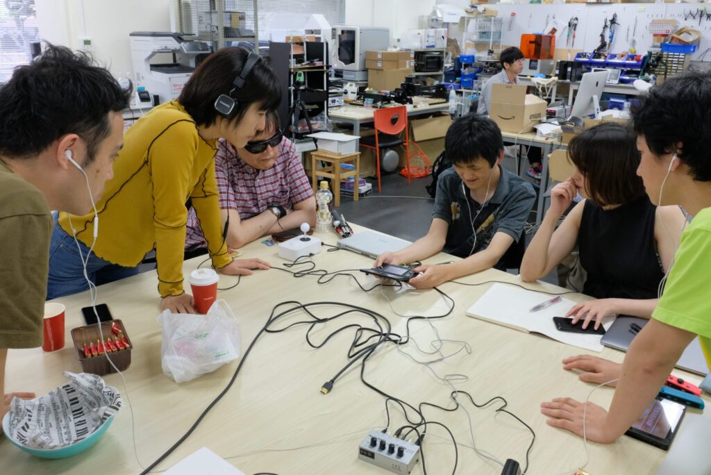 6人の男女がテーブルを囲んで電子機器を操作している。テーブルの上にはケーブルやデバイスが広がっており、皆がそれぞれイヤホンやヘッドホンを着用している。1人の男性がスマートフォンを操作しており、他の人々がそれを興味深く見つめている。背景には多くの機材や箱が積まれた作業スペースが見える。