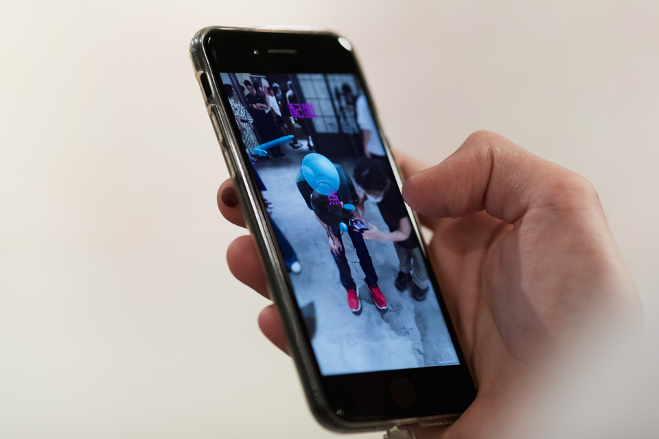手に持ったスマートフォンの画面に、拡張現実(AR)技術を使っている様子が映し出されている。画面には青い球体とその背後に立つ人々が見える。スマートフォンを持つ手がしっかりと画面を支えている。