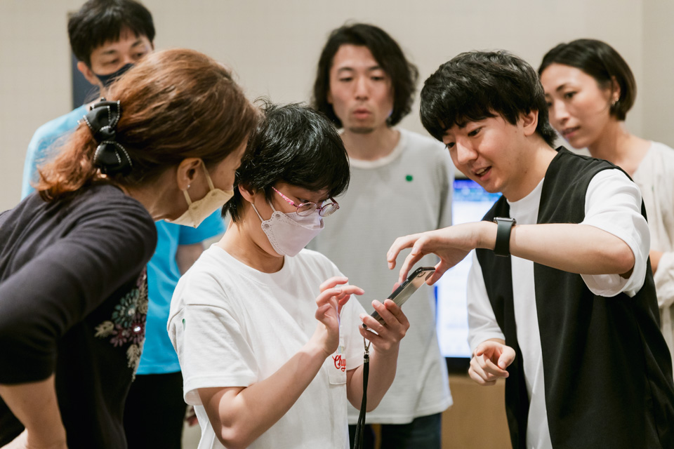 数名の男女が集まり、マスクを着けた女性がスマートフォンを操作しているところを見ている。右側の男性が女性に操作方法を教えているようで、その他の人々も興味深く画面を見つめている。背景には別の人々も写っており、皆が関心を持っている様子が伺える。