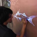 黒い髪の男性が、壁に白い魚の絵を描いている。男性はメガネをかけていて、絵に集中している。背景には魚のシルエットがいくつか見える。