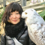 女性が大きな白いフクロウと一緒に写っている。女性は黒いコートを着て、微笑んでカメラを見ている。背景には緑の植物が見える。