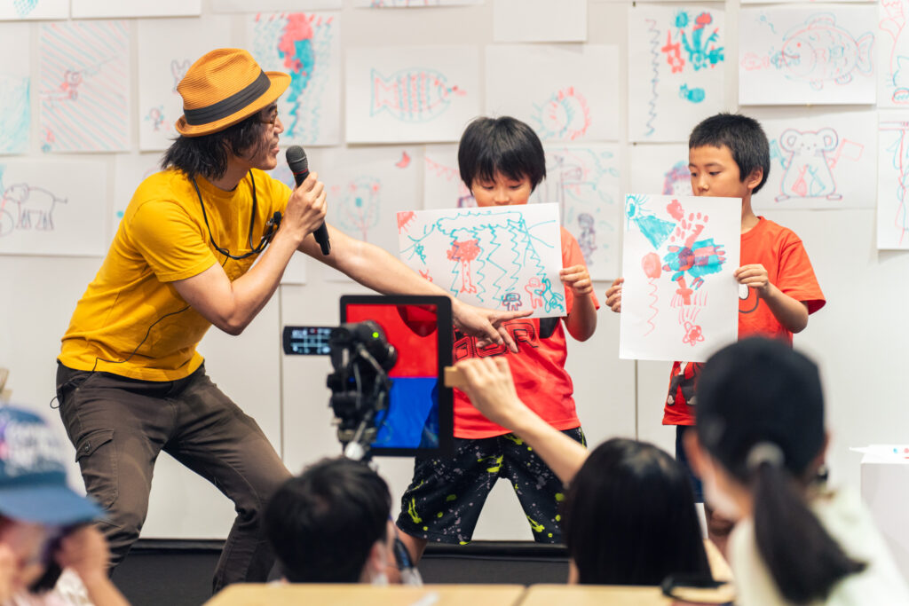 アニメーションのワークショップで、黄色いシャツを着た男性がマイクを持ち、2人の子供が自分たちの描いた絵を見せている。後ろの壁には多くのカラフルな絵が貼られていて、観客が前方でその様子を見守っている。