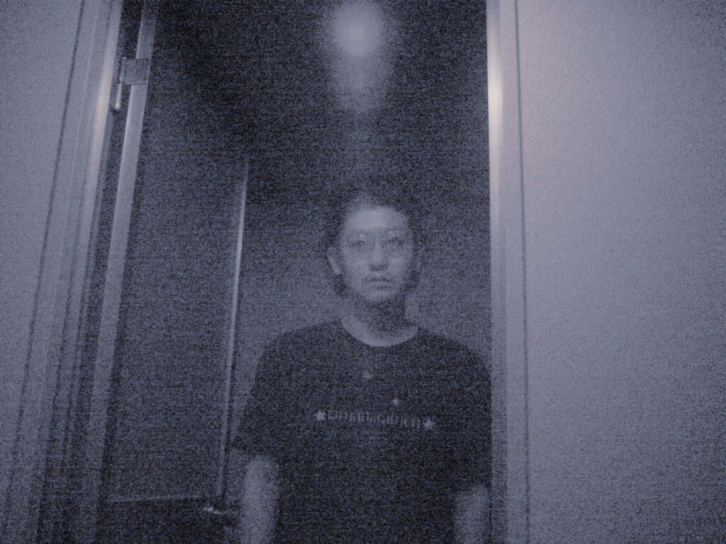 ざらついたモノクロ画面に、ドア枠の前で薄いガラス越しにこちらを直視する男性の写真