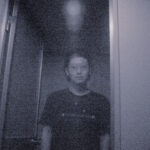 ざらついたモノクロ画面に、ドア枠の前で薄いガラス越しにこちらを直視する男性の写真