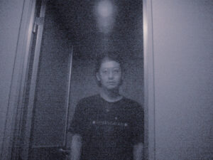 ざらついたモノクロ画面に、ドア枠の前で薄いガラス越しにこちらを直視する男性の写真