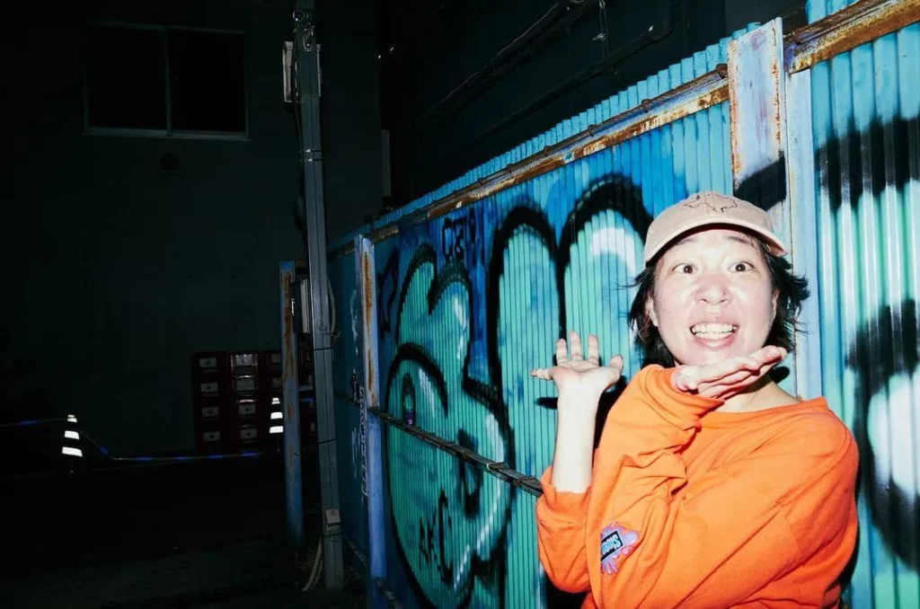 グラフィティの描かれた青い金属壁を背に、ベージュのキャップとオレンジの長袖シャツを着て、両手を頬の下に添え、満面の笑みでカメラを見つめている女性の写真