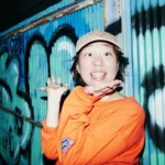グラフィティの描かれた青い金属壁を背に、ベージュのキャップとオレンジの長袖シャツを着て、両手を頬の下に添え、満面の笑みでカメラを見つめている女性の写真
