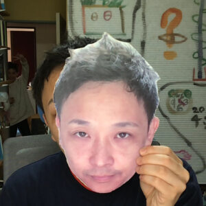 きな男性の顔をかたどった紙製マスクを手に持ち、椅子に座っている男性の写真
