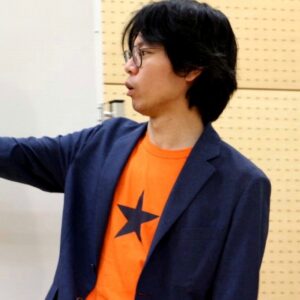 星柄オレンジTシャツにネイビーブレザーを羽織った眼鏡をかけた男性