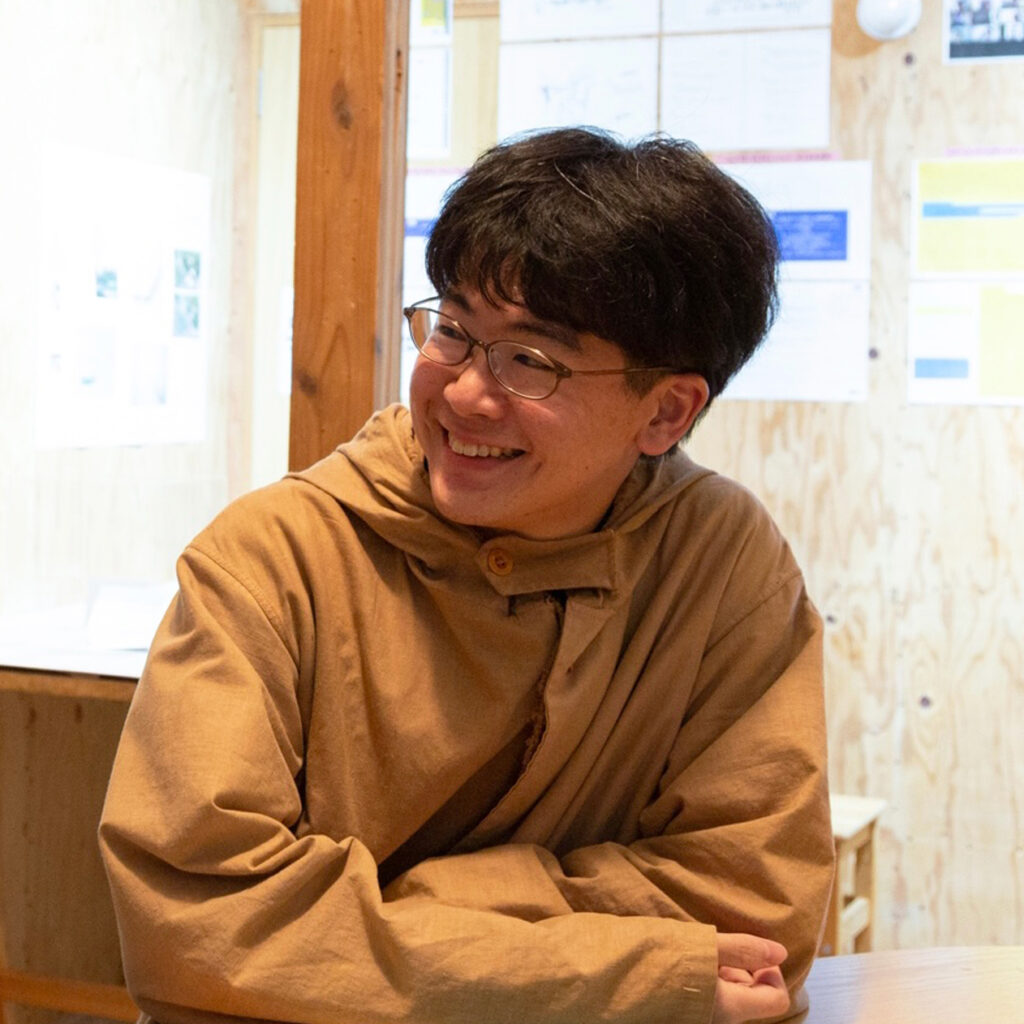 キャラメルブラウンのフード付きジャケットを羽織り、木目の壁が並ぶ室内のテーブルに肘をついて座っているメガネの男性