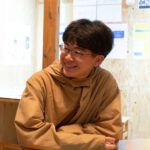 キャラメルブラウンのフード付きジャケットを羽織り、木目の壁が並ぶ室内のテーブルに肘をついて座っているメガネの男性