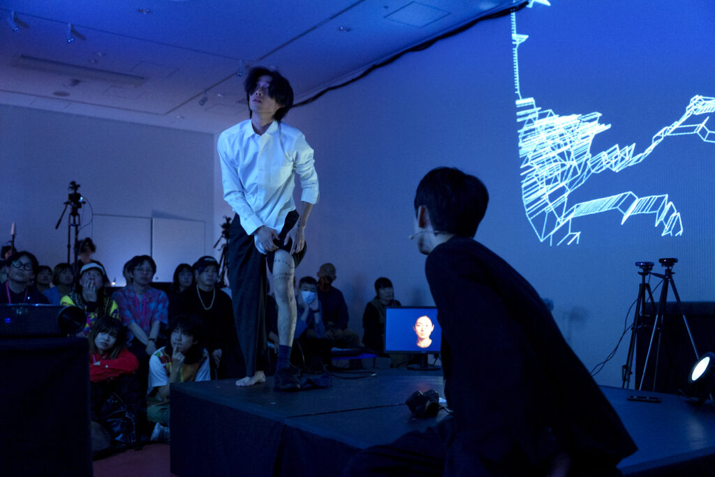 青い照明の中、白いシャツを着た演者がステージ上でズボンを少し上げ、脚を見せながらパフォーマンスをしている様子の写真