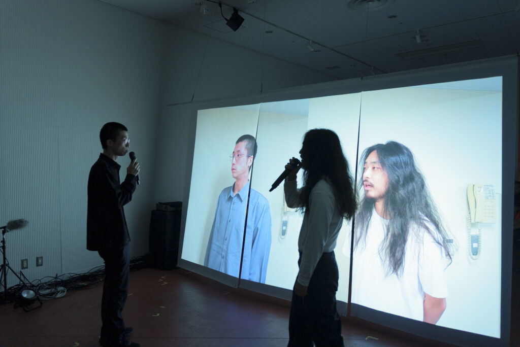 映像が投影されている透過型のスクリーンの前で、映像に登場している2名の演者がマイクを持って話している様子の写真
