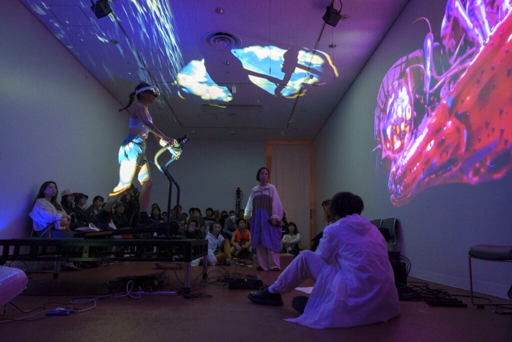 暗い室内の中央で3名の演者がパフォーマンスを行っている様子の写真。ひとりは、エアロバイクをこぎ、もうひとりは床に座り、最後の一人は中央に立っている。壁や天井には鮮やかな映像が投影されている。