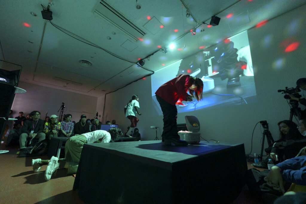 暗い照明とカラフルな光の中、室内ステージで複数の出演者が身体を使ったパフォーマンスを行っている様子の写真