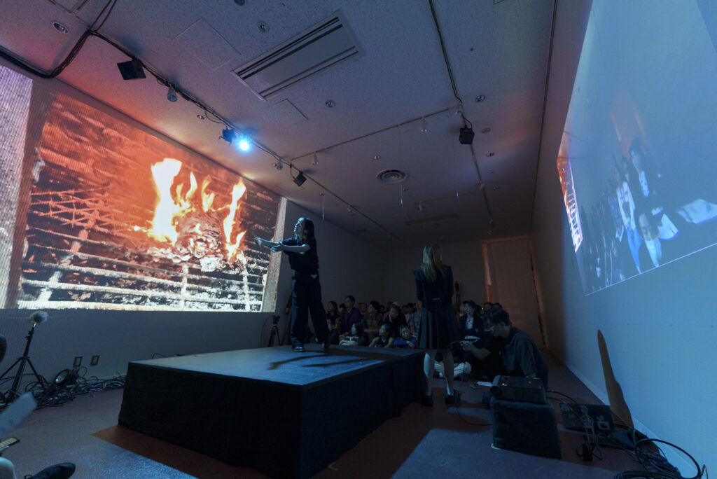 暗い室内で、ステージ上の人物が手を広げてパフォーマンスをしている様子の写真。壁のスクリーンには炎が燃える映像と観客の姿が投影されている。