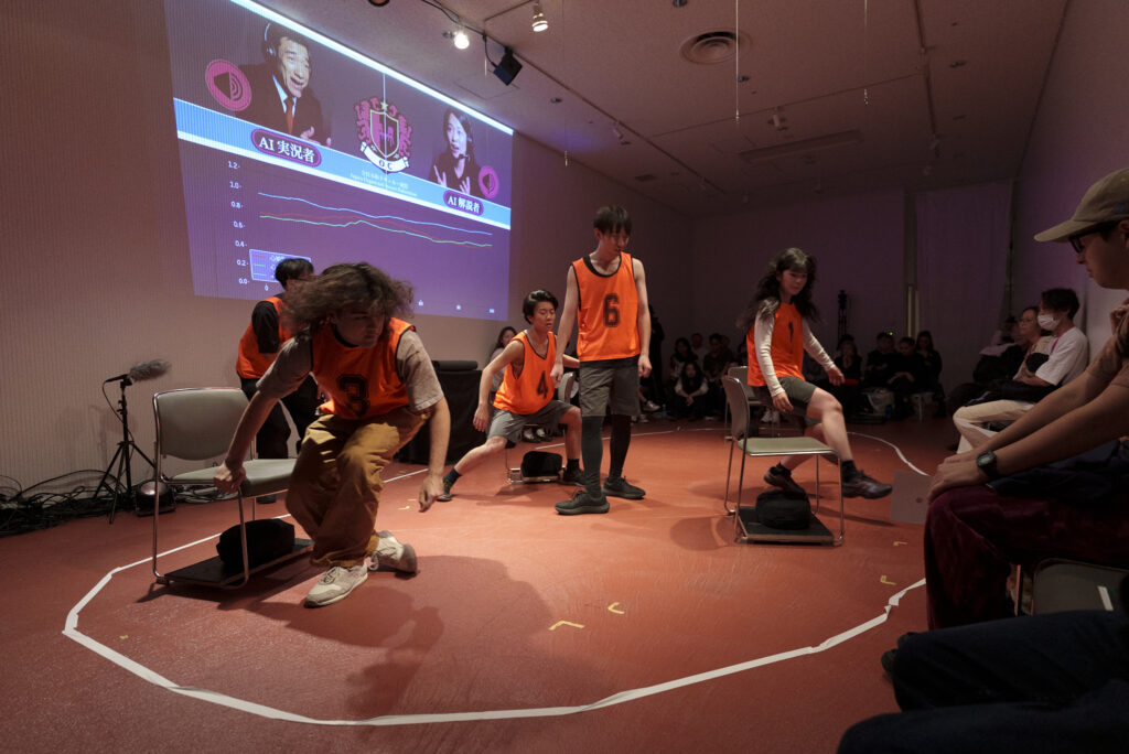 ステージでオレンジ色のビブスを着た数人の参加者4人が椅子を使ってパフォーマンスを行っている様子の写真