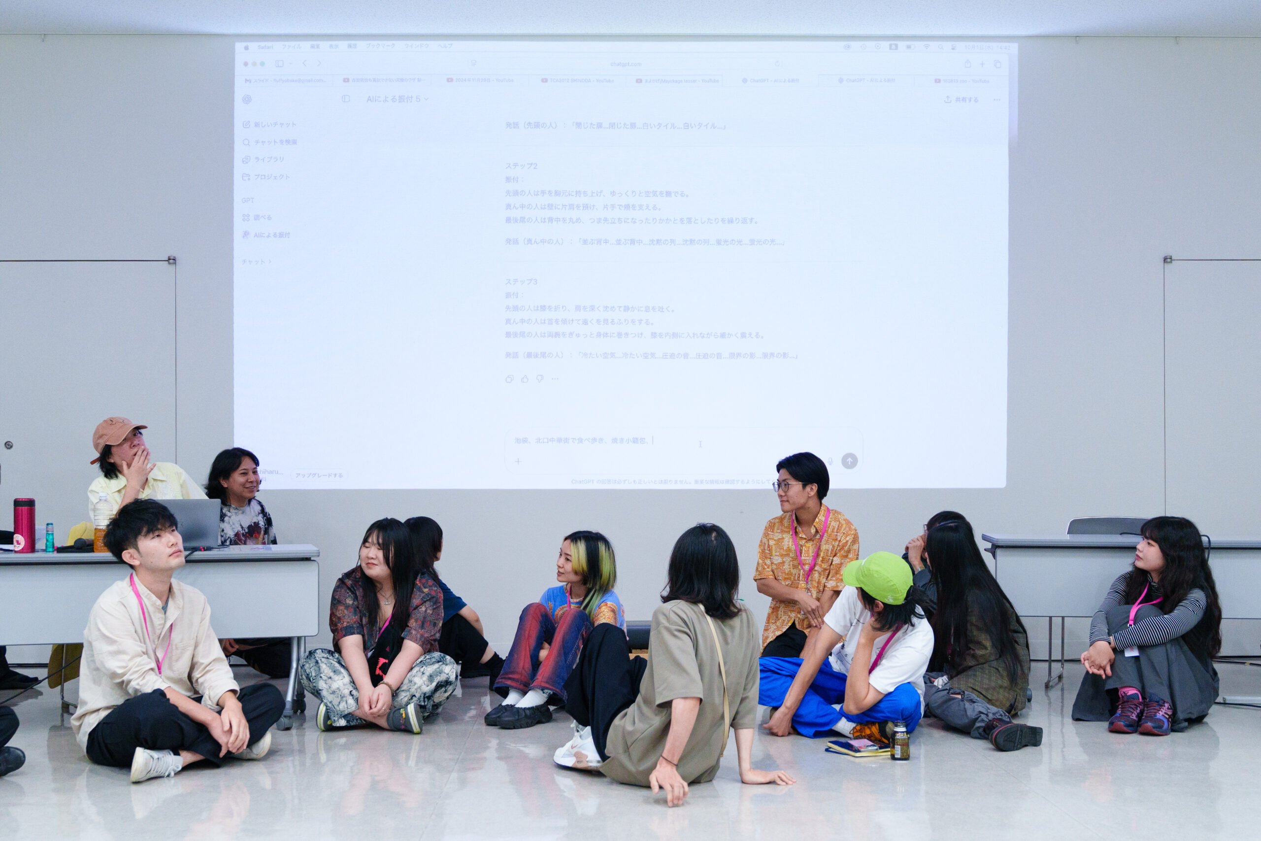 参加者が床に座り、大きなスクリーンに投影されたChatgptが生成した振り付けを確認している様子の写真