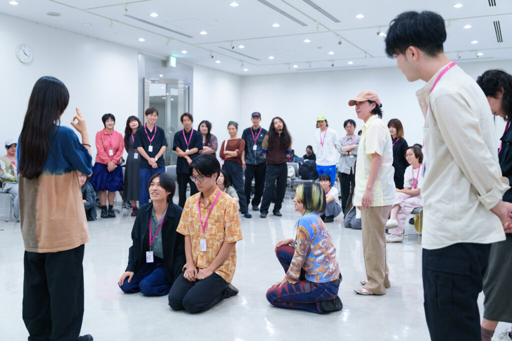 篠田さんのレクチャーでの一場面の写真。参加者たちが輪になって集まり、その中央では数人が床にひざまずいたり座ったりして、向かい合いながらやりとりをしている。即興で篠田さんが演出したシーンが展開されている。