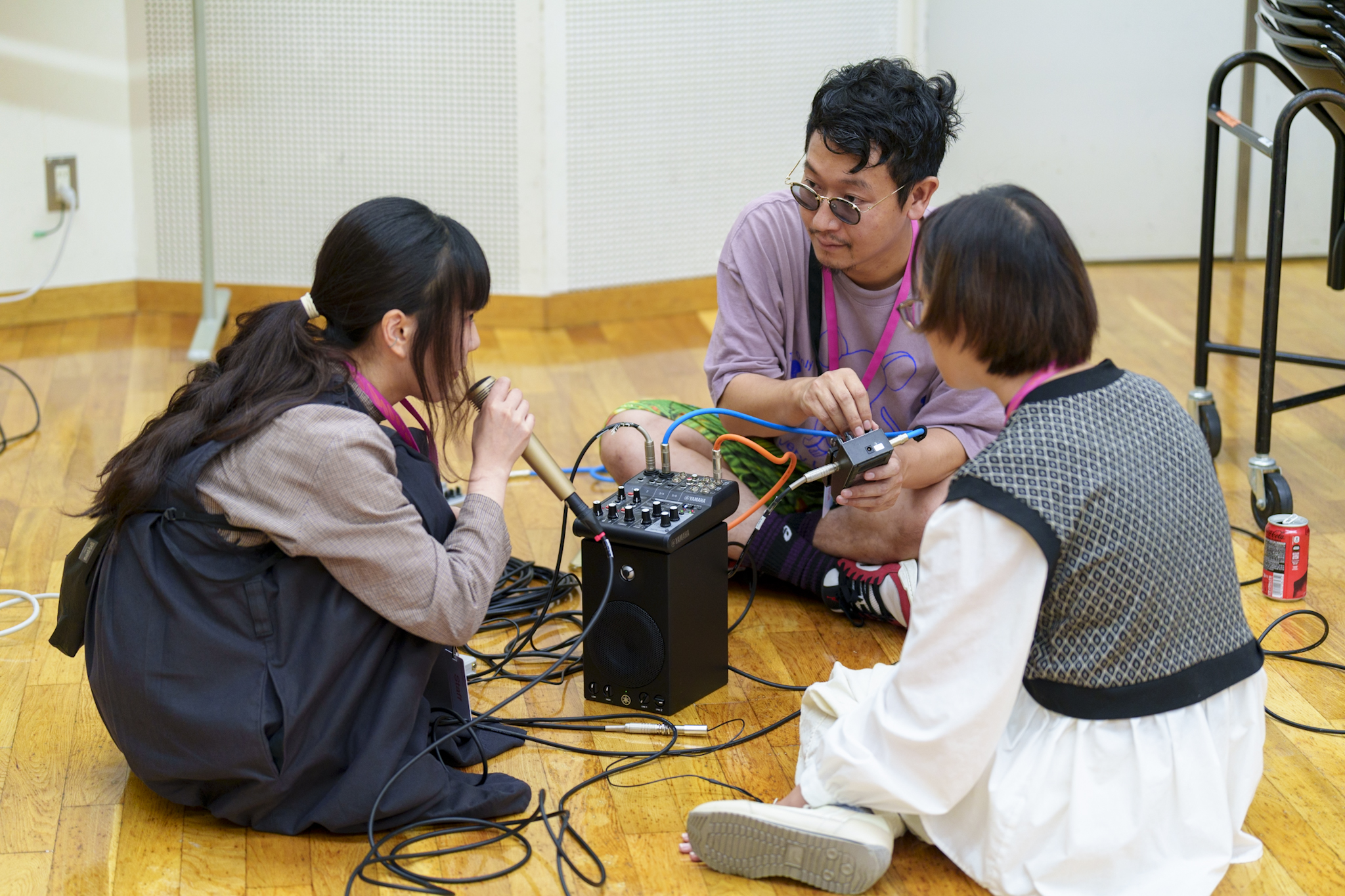 二人の参加者とファシリテーターが床に座り、音響機器を囲んで作業している様子の写真。1人はマイクに向かって話し、もう1人はケーブルを操作し、もう1人はその様子を見ている。