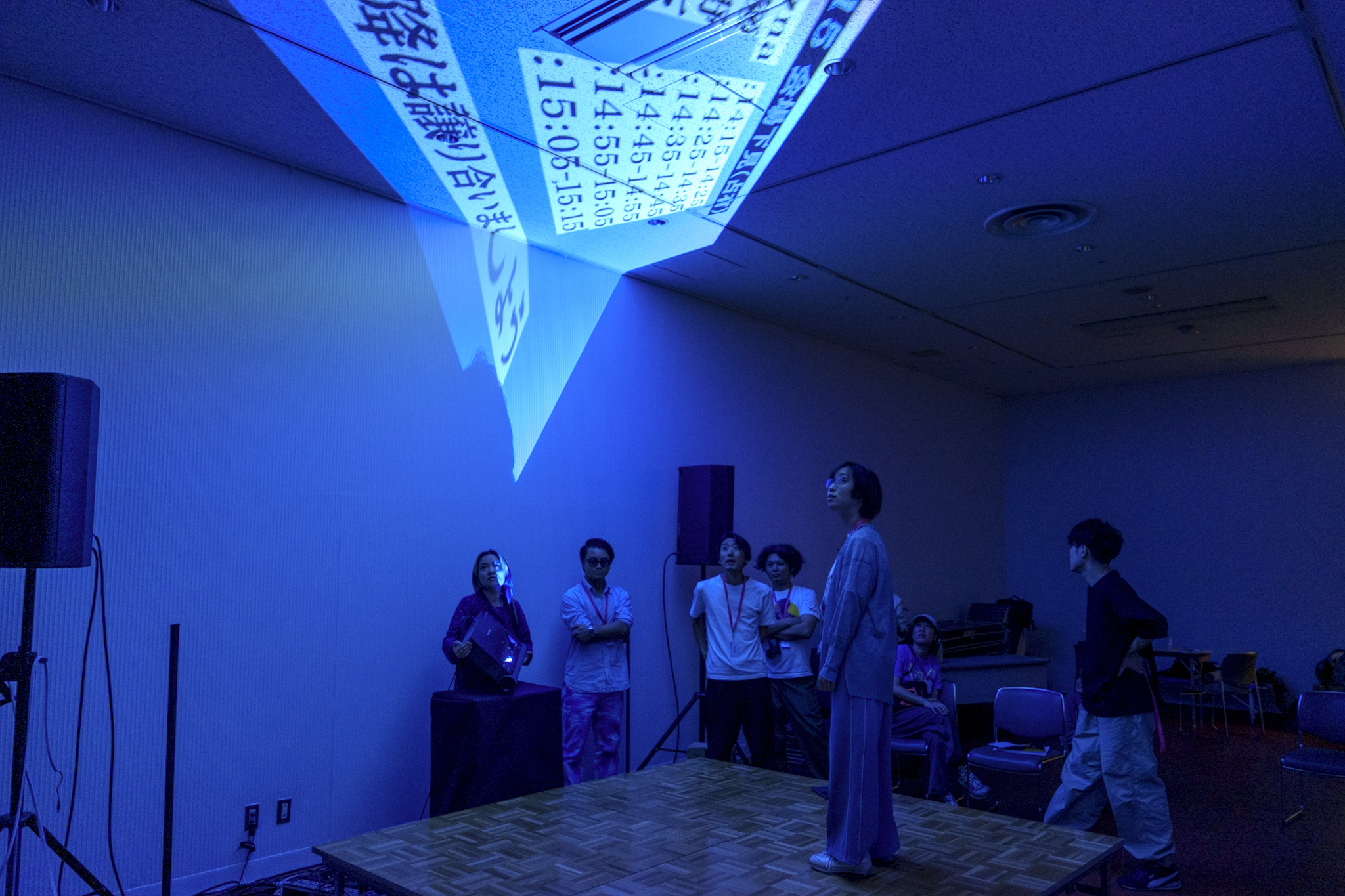 暗い室内で青い照明に照らされた空間のなかで、天井近くに文字や映像が投影されており、数人の参加者がそれを見上げている様子の写真